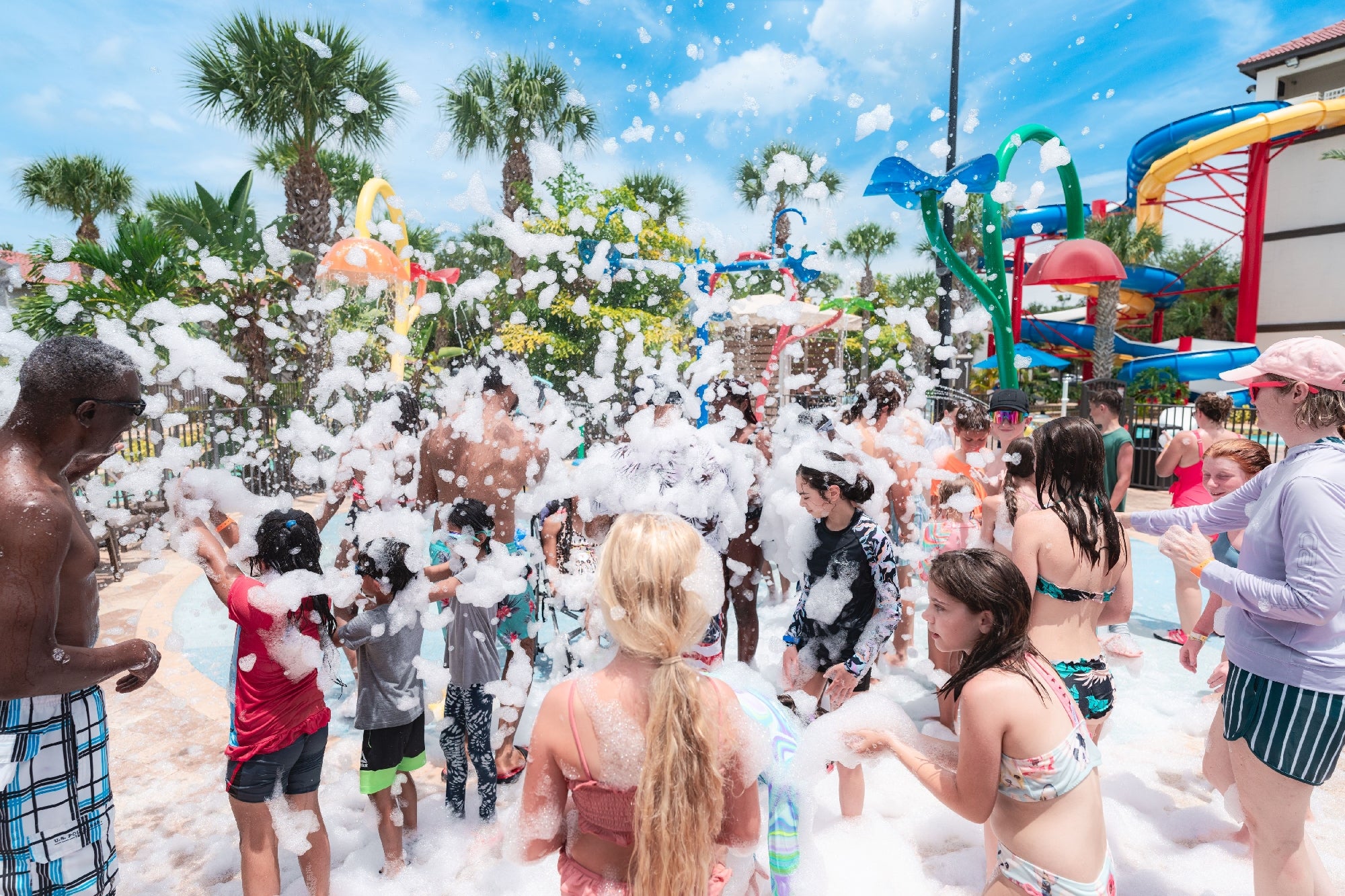 A group of people, including children and adults in swimwear, play in foam at an outdoor water park with palm trees and colorful slides in the background.