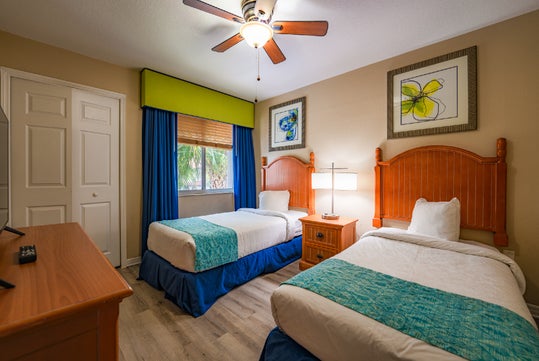 A hotel room with two single beds, teal bed runners, wooden headboards, a nightstand with a lamp, a ceiling fan, and framed artwork on beige walls.