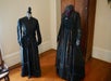 Two antique black dresses are displayed on headless mannequins in a room with wooden floors and a wooden wardrobe in the background.