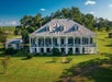 Aerial view looking down on the front of a large white two story plantation home with a shed to the left and trees behind it on a sunny day.