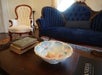 Close up a dark wooden coffee table with old books and an ornate bowl sitting on it with a deep blue vintage couch and a cream vintage chair behind it.