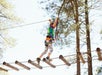 A person wearing a harness crosses a rope bridge made of logs in an outdoor adventure park surrounded by tall trees.