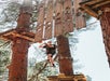 A person wearing a harness climbs a vertical obstacle on an outdoor ropes course among tall trees.