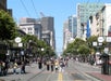A busy city street scene with people crossing, surrounded by tall buildings under a clear blue sky.