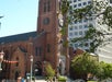 A red brick church with a clock tower stands next to a modern white office building. Trees and street signs are visible in the foreground.