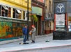 Two men stand on a city sidewalk; one plays a guitar and sings. Colorful murals and a bookstore are in the background.
