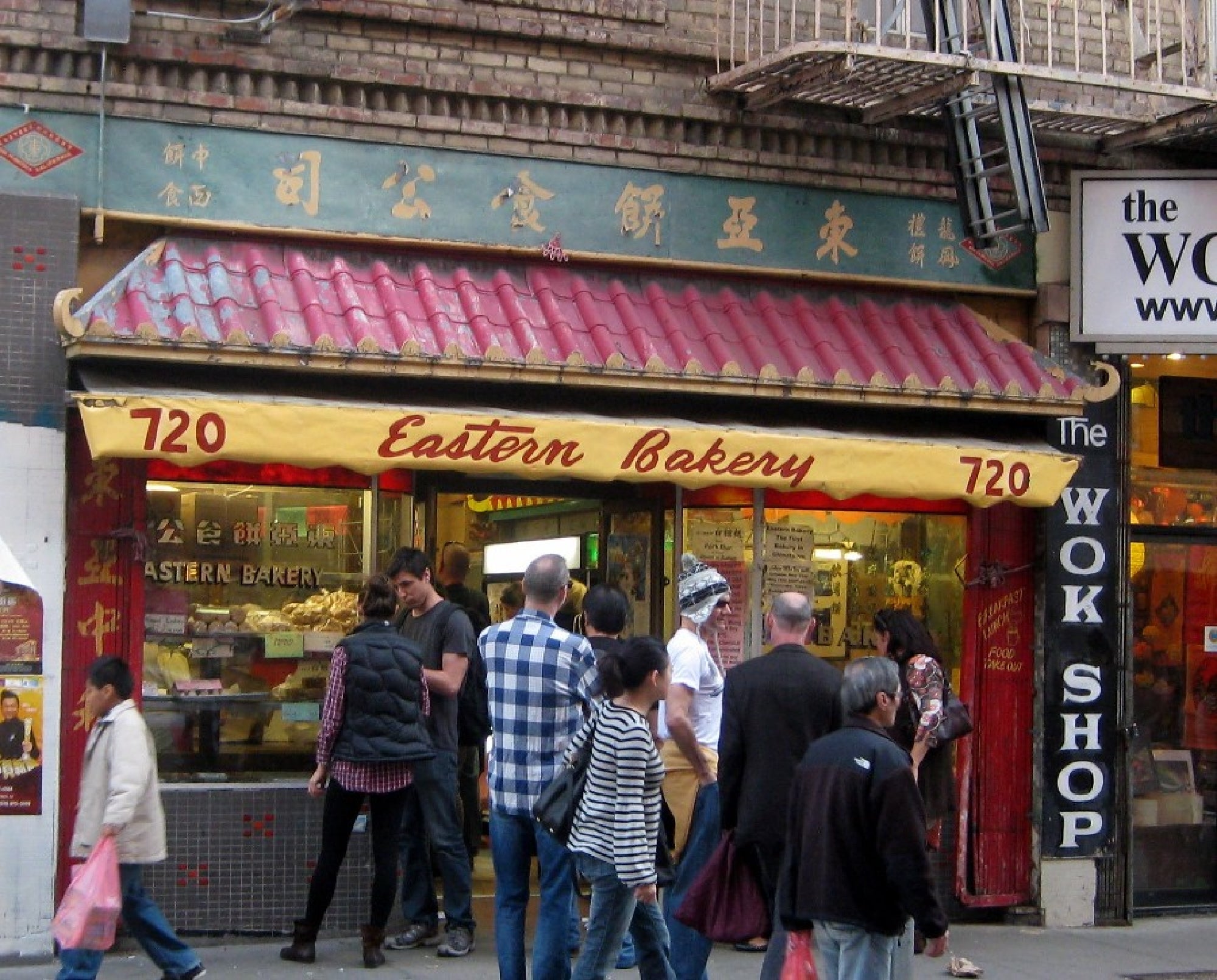 People standing outside Eastern Bakery with a yellow awning, located next to "The Wok Shop." The bakery has a red-tiled roof and displays sweets in the window.