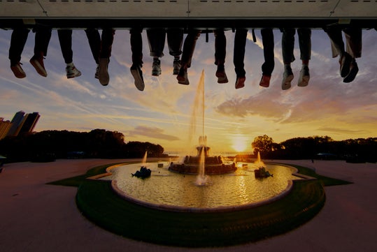 A row of legs and feet dangle from above, silhouetted against a sunset over a large fountain in a landscaped park.