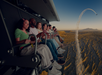 A group of people sit in seats on a simulated flying ride above a desert landscape, watching a jet perform an aerial maneuver in the sky.