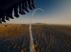 People's feet hang from an aircraft as they watch a stunt plane creating a smoke trail loop over a rugged desert landscape at sunset.