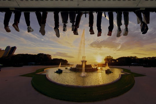People's legs and feet hang from above, silhouetted against a sunset sky, with a large fountain and landscaped park in the background.