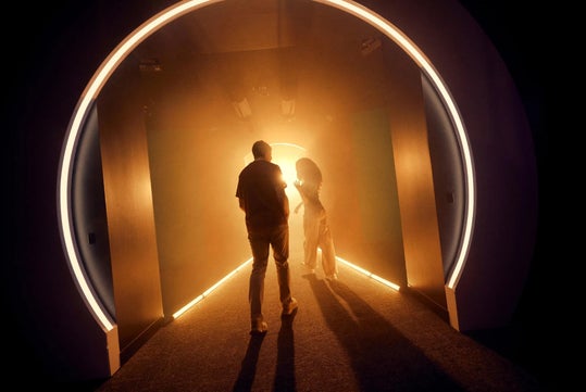 Two people walk through a circular, illuminated tunnel with warm light and shadows surrounding them.