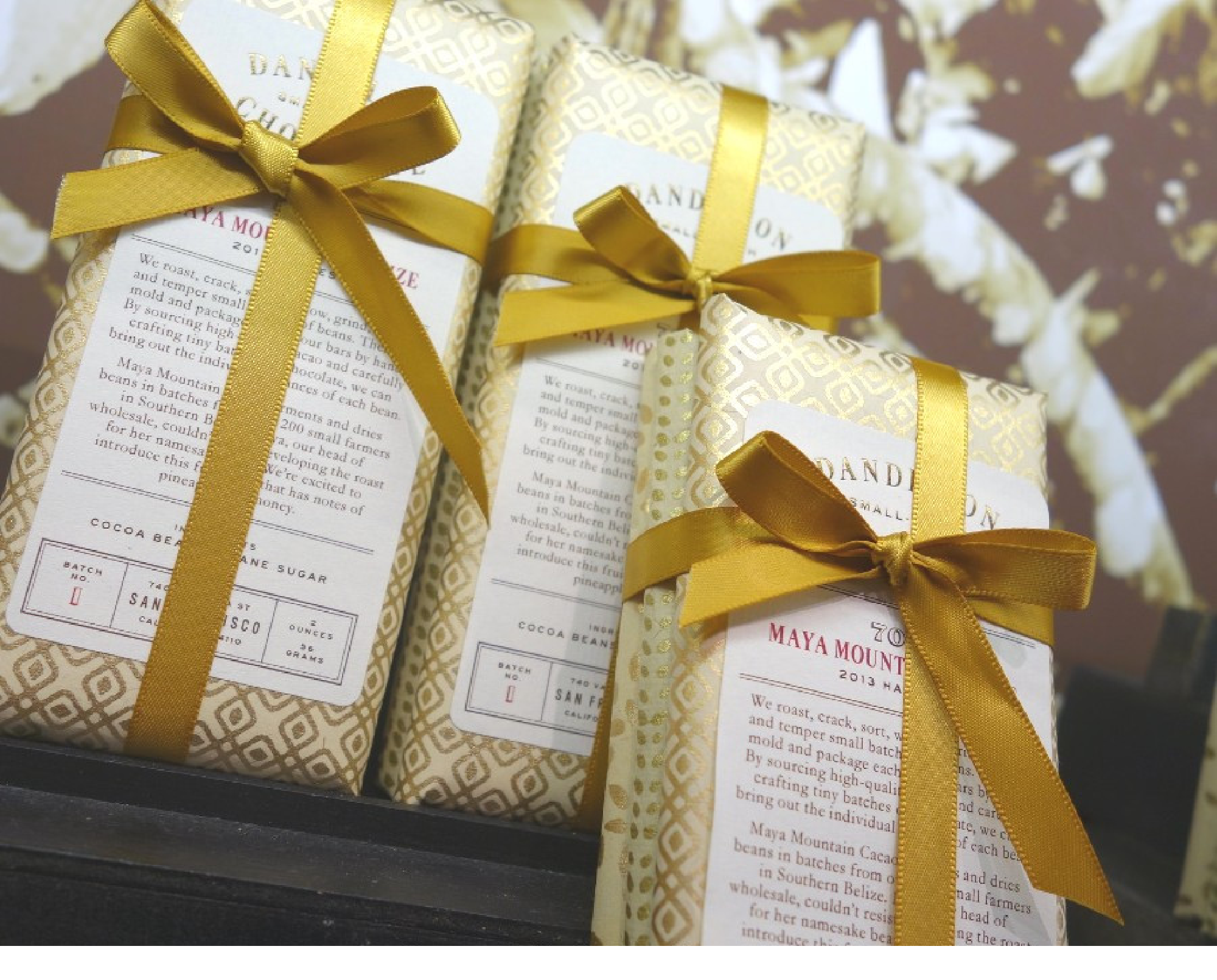 Three decorative chocolate bars wrapped in gold-patterned paper with gold ribbon bows. The packaging reads "Dandelion" and "Maya Mountain.