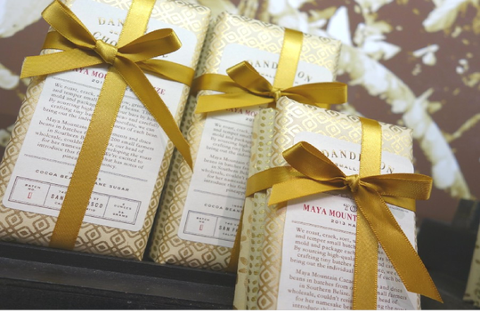 Three decorative chocolate bars wrapped in gold-patterned paper with gold ribbon bows. The packaging reads "Dandelion" and "Maya Mountain.