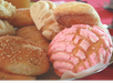 A close-up of a plate with various sweet breads, including a pink concha, a sugar-coated pastry, and a sesame seed roll, on a red tablecloth.