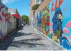 Colorful murals line a narrow alley with a vibrant blue sky above. A couple of people walk in the distance, surrounded by graffiti-covered walls.