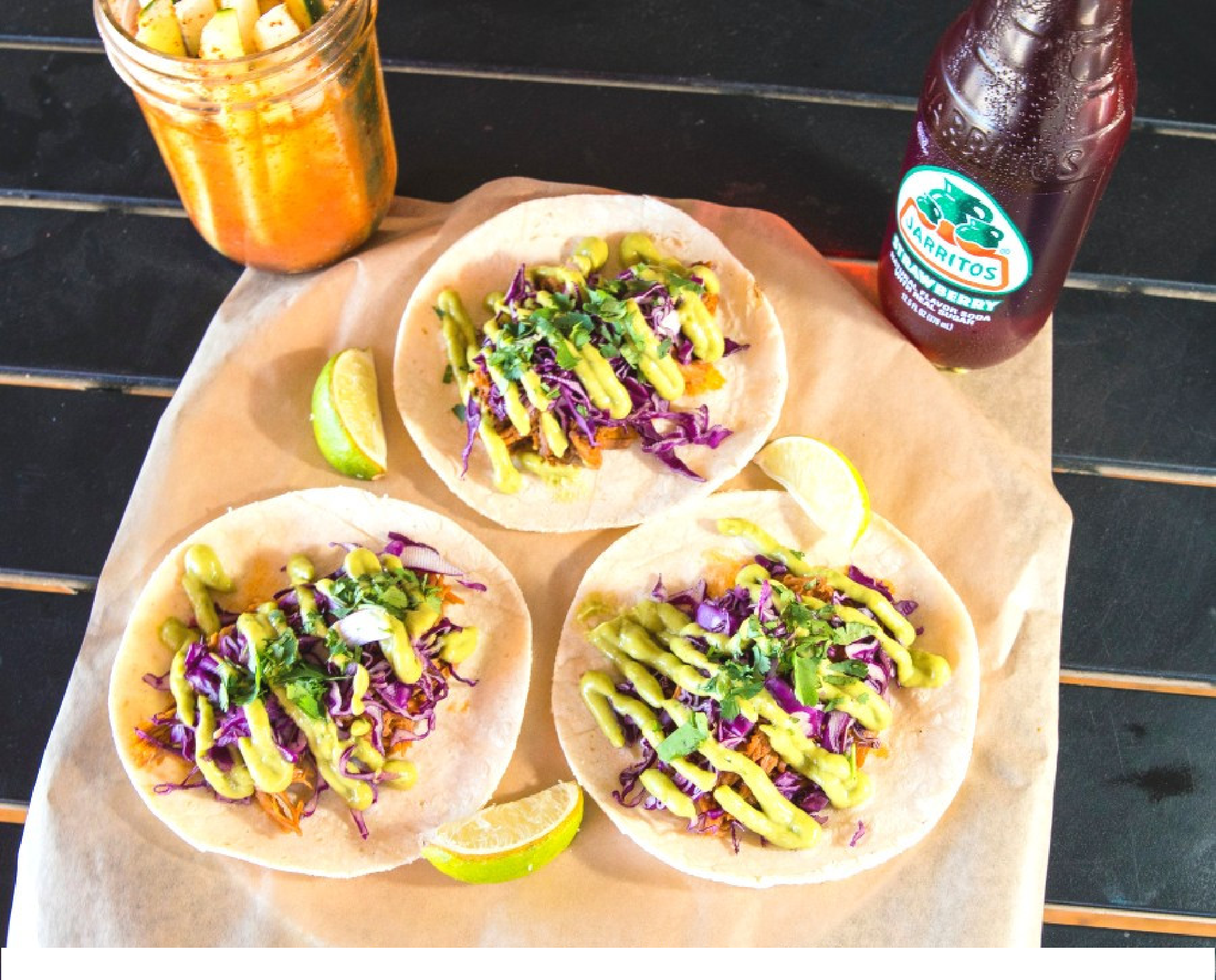 Three tacos with shredded meat, purple cabbage, greens, and sauce on tortillas, next to lime wedges, a jar of red sauce, and a bottle of soda on a wooden surface.