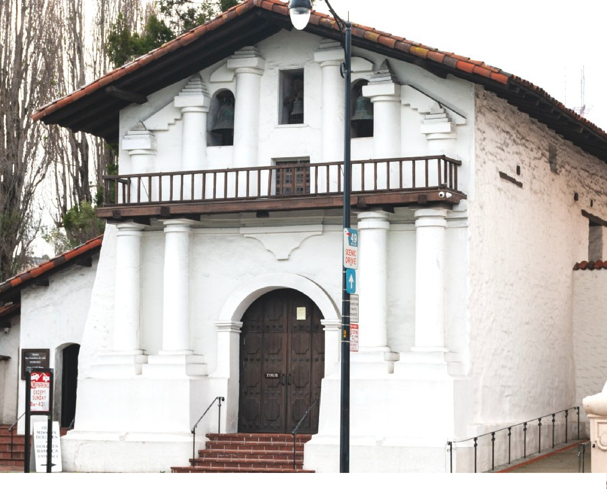 A historic white adobe building with a wooden balcony and arched entrance, framed by slender pillars, stands next to a "No Parking" sign and a streetlight.