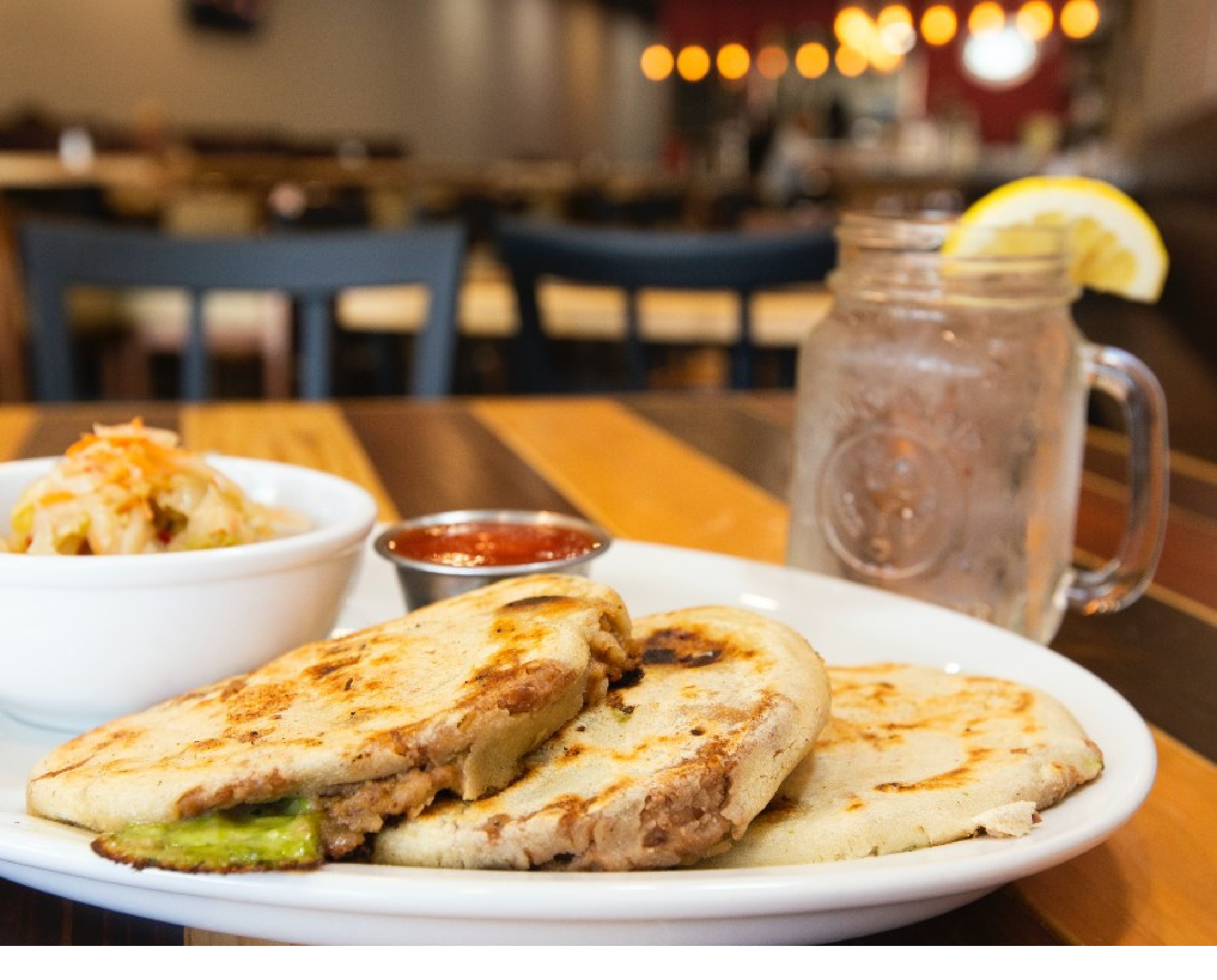 Plate of pupusas with a side of salsa and coleslaw on a wooden table. A mason jar with a lemon wedge and a drink is in the background.
