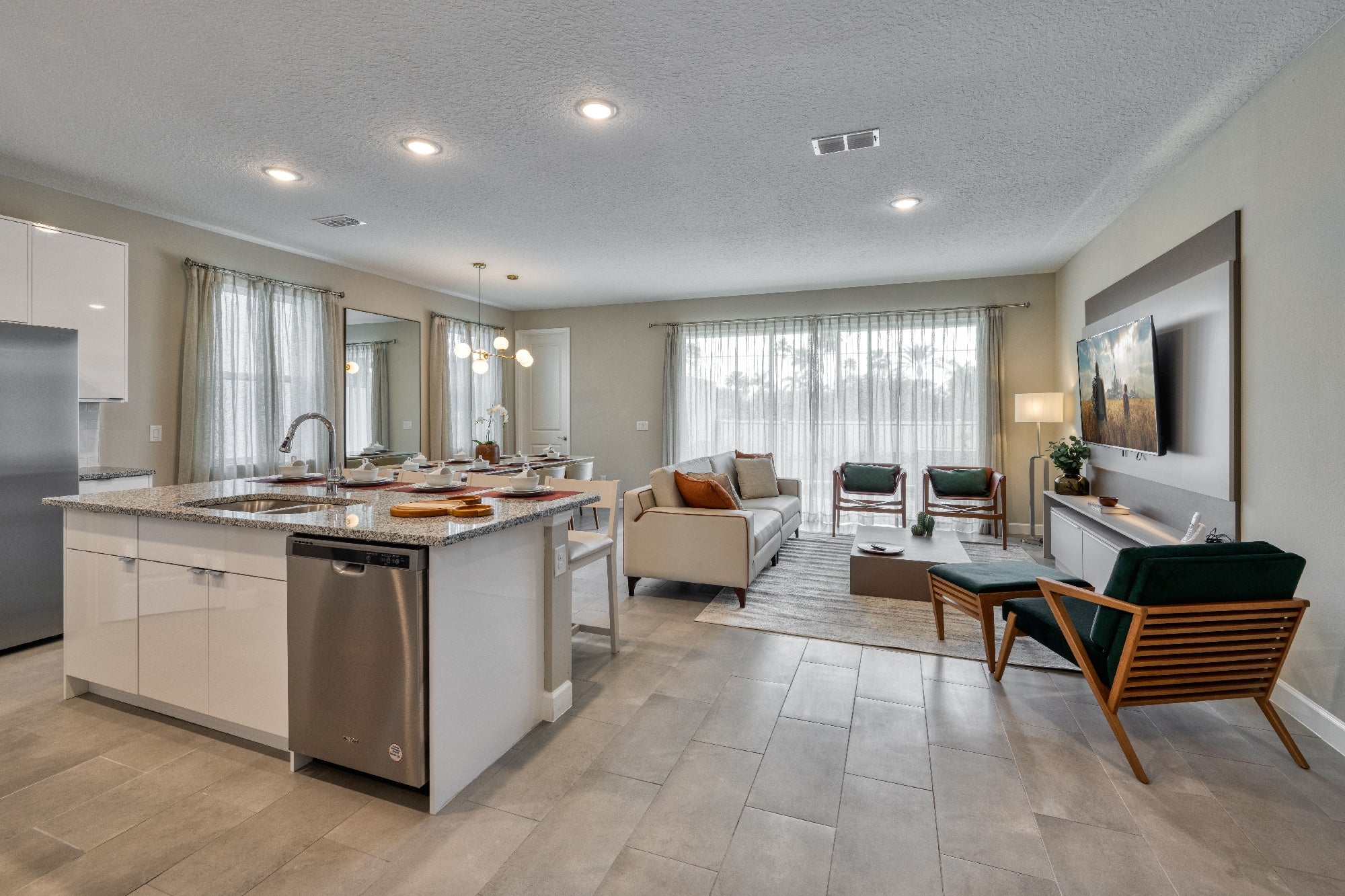 Modern open-concept living room and kitchen with an island, white cabinets, dining table, sofa, chairs, TV on the wall, and large windows with sheer curtains.