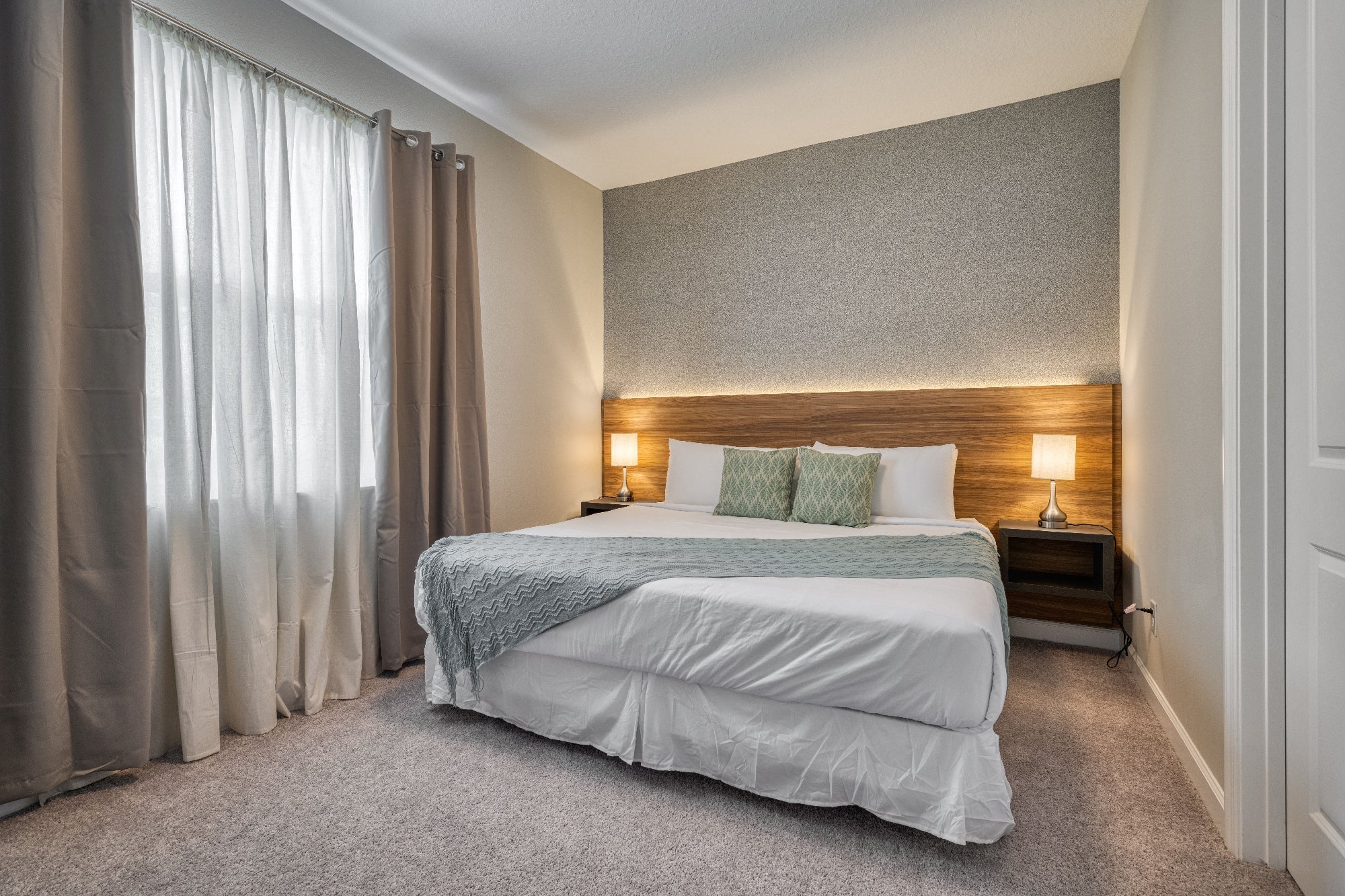Bedroom with a large bed, two bedside tables with lamps, curtain-covered window, and gray carpet. The bed has white linens and a gray throw, with a textured wall behind the headboard.