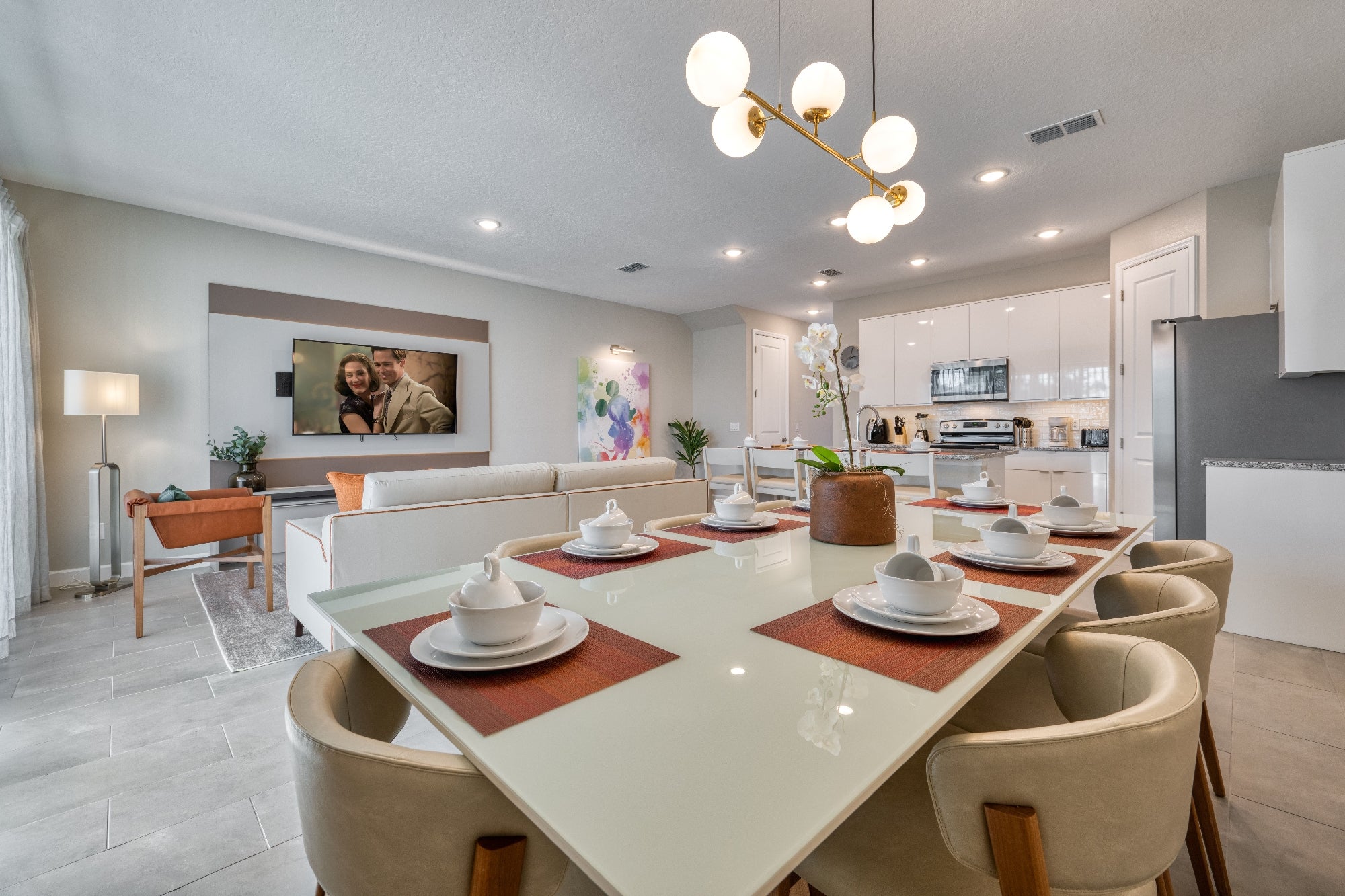 Modern open-concept living area with a glass dining table set for six, white sectional sofa, artwork, and a wall-mounted TV across from the kitchen. Ceiling light fixture is prominent.