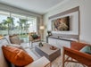 Modern living room with a large TV, two armchairs, a sofa, and a coffee table. Sliding glass doors open to a patio with a view of the pool and garden.