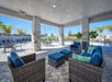 Outdoor lounge area with wicker furniture and blue cushions under a covered patio. Beyond the patio, there is a swimming pool surrounded by lounge chairs and palm trees.