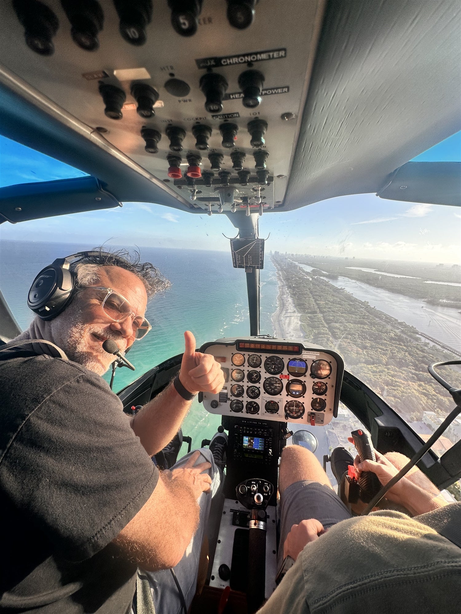 A person with a headset gives a thumbs-up while sitting in the cockpit of a helicopter flying over a coastal area.