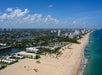 Aerial view of a wide sandy beach lined with resorts and high-rise buildings, bordered by the ocean on one side and a cityscape with waterways on the other under a partly cloudy sky.
