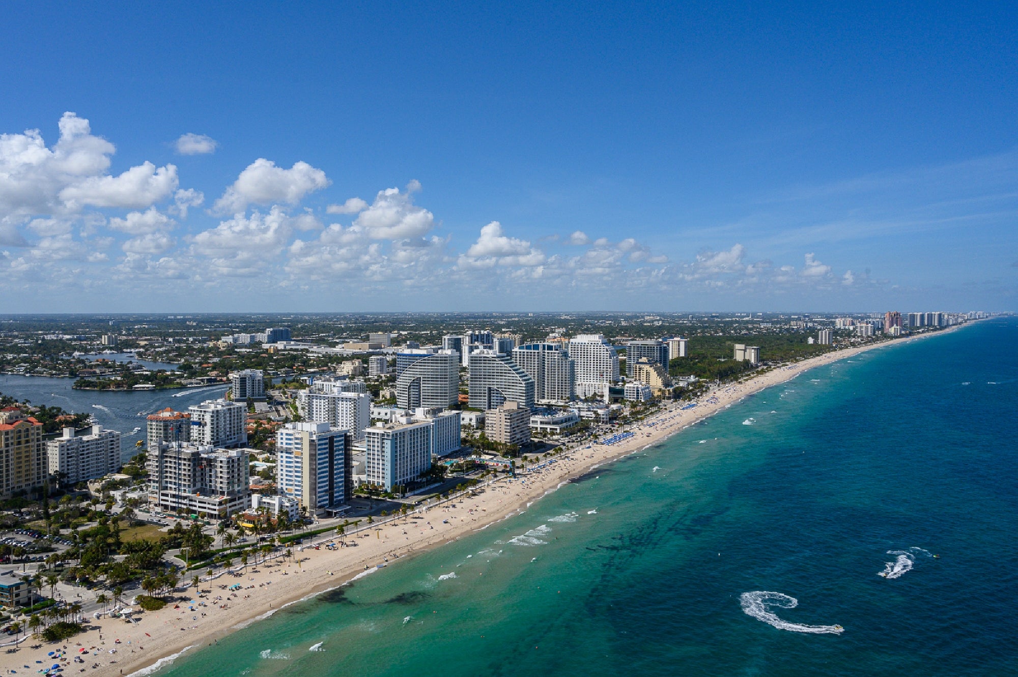 Aerial view of a coastline with high-rise hotels and buildings along a sandy beach, with turquoise ocean water and blue sky with scattered clouds.