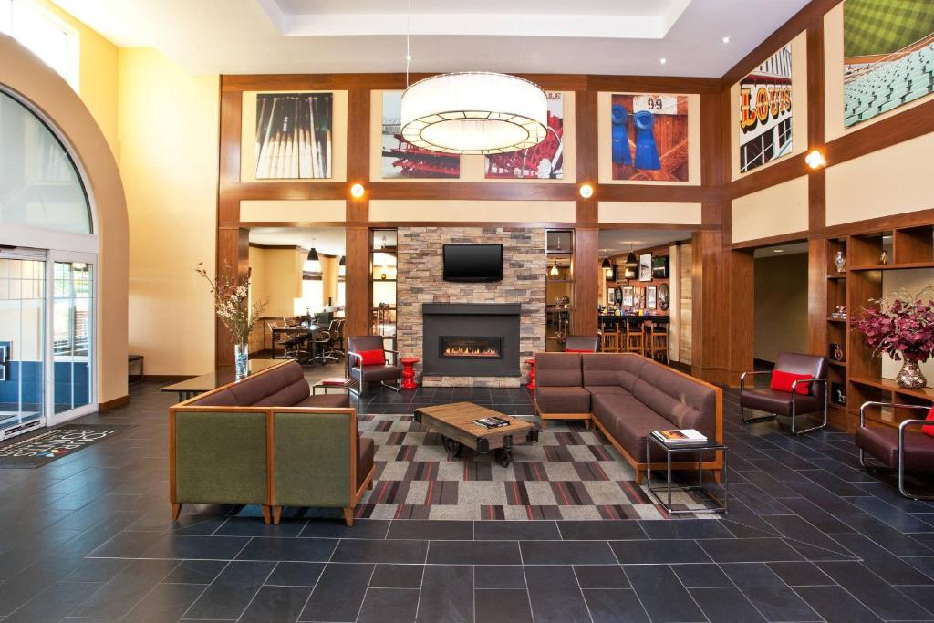Hotel lobby with modern decor, featuring a central seating area, stone fireplace, mounted TV, and artwork on the walls. Large windows and a light fixture illuminate the space.