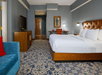 Hotel room with a large bed, blue-patterned carpet, blue sofa with red pillow, TV on a wooden stand, and a desk with a chair and lamp near the entry door.