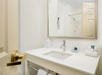 Bathroom with a white sink, chrome faucet, and a large mirror above. Towel rack below the countertop, toiletry items, and a tissue box are on the counter. Shower visible in the mirror.