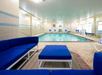 Indoor swimming pool with blue cushioned seating. Bright lighting reflects off the water, surrounded by tiled walls and windows. Safety equipment and signs are visible on the walls.