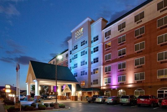 Exterior view of a multi-story hotel at dusk, with a lit entrance, parked cars in the foreground, and a sunset sky in the background.