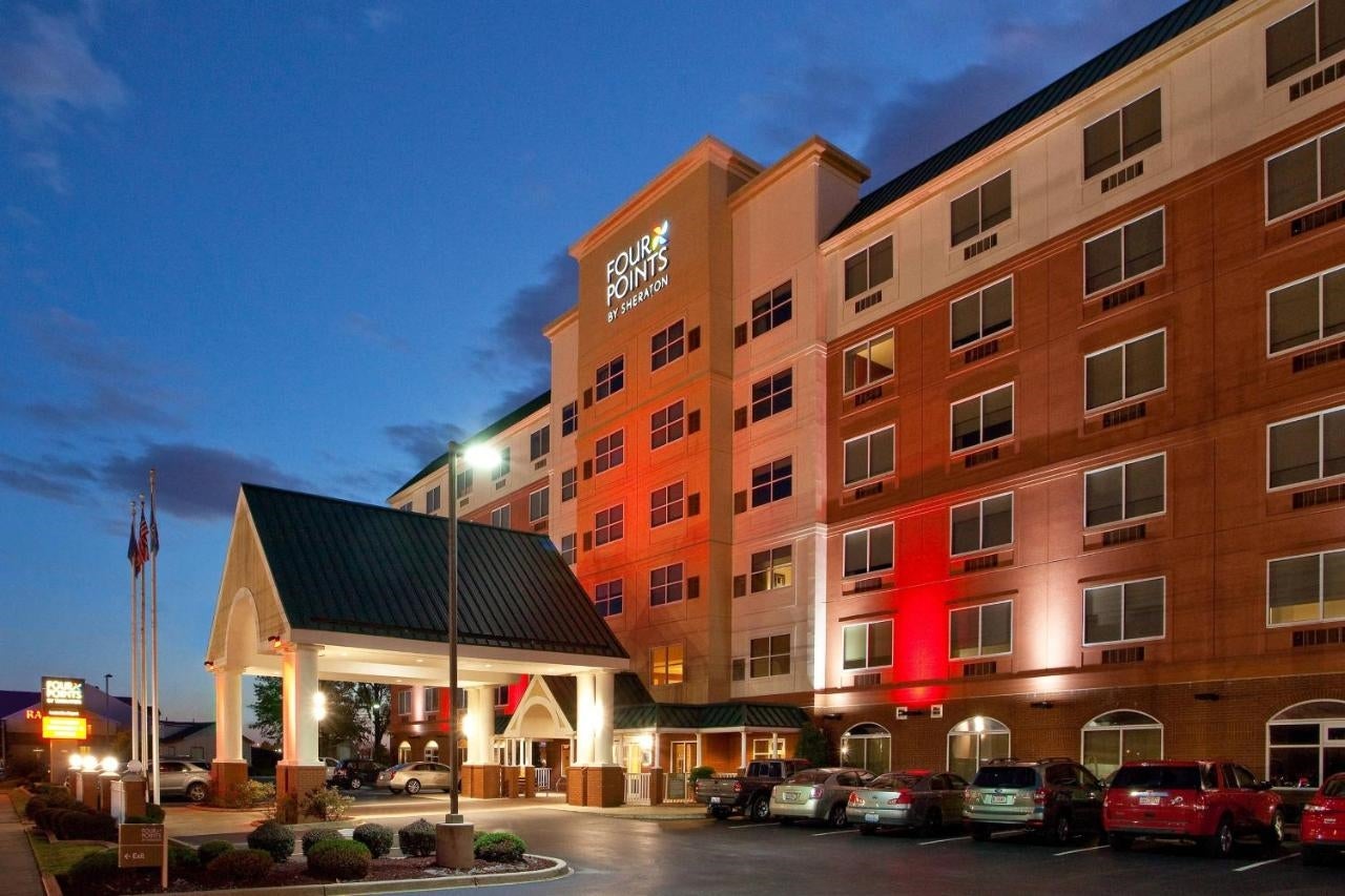 Exterior view of a mid-rise hotel with a covered entrance and cars parked out front, illuminated by exterior lights at dusk.