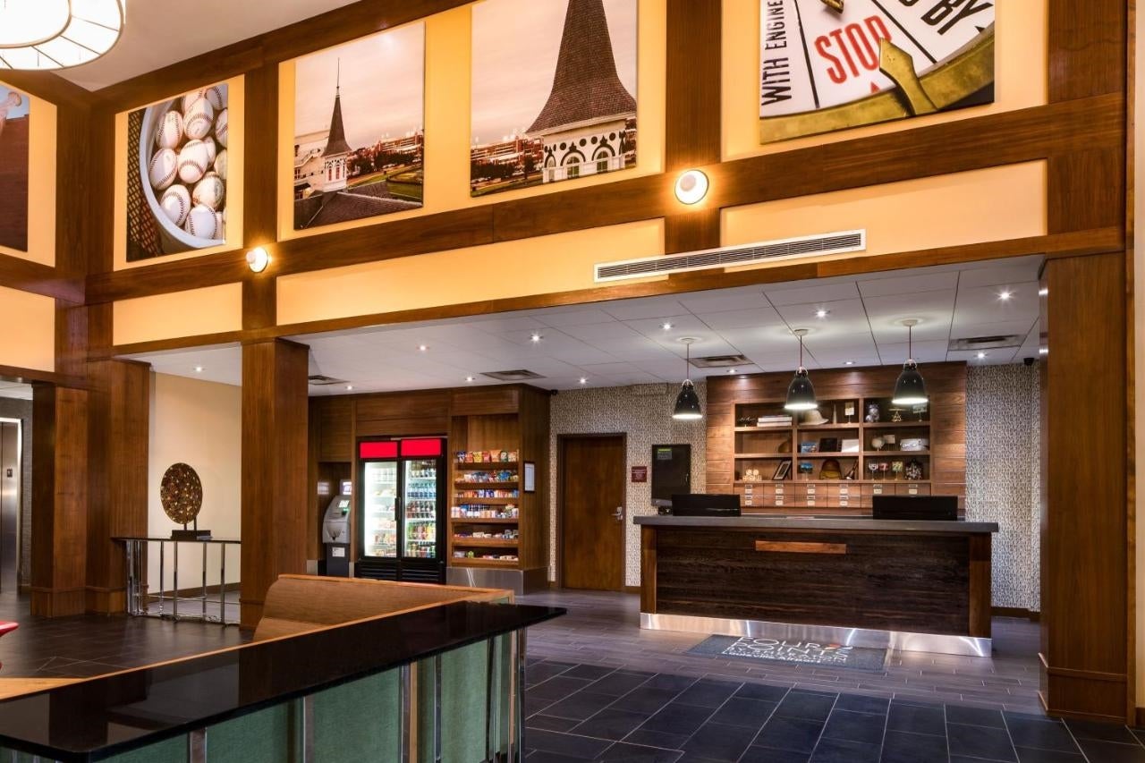 Hotel lobby featuring a modern reception desk, a snack area with vending machines, and decorative wood paneling. Above, large photos adorn the walls.