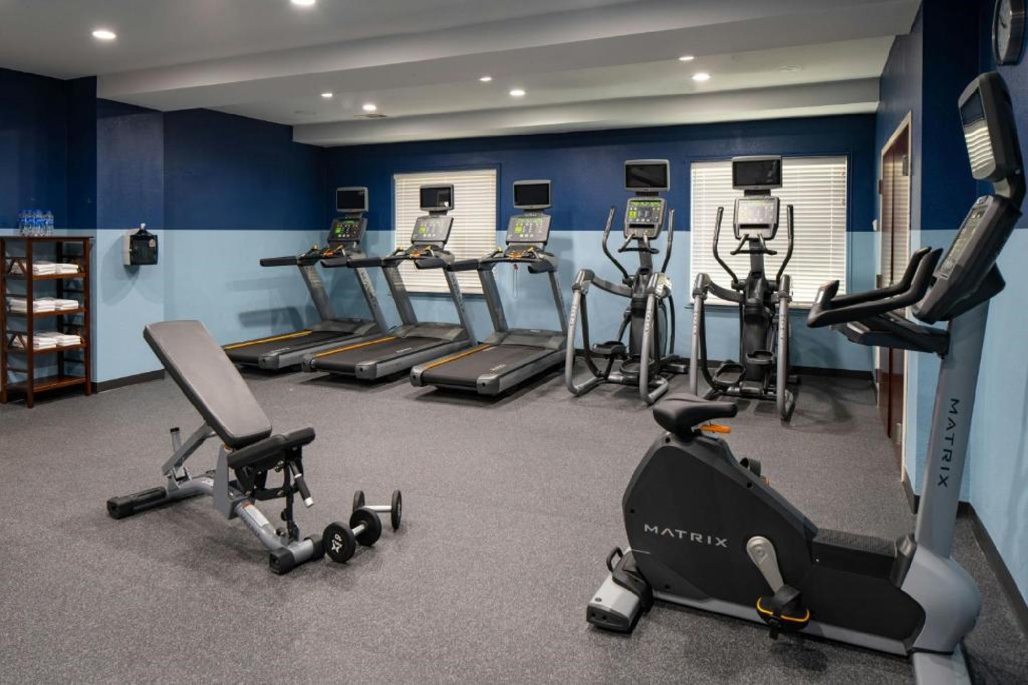 A fitness room with treadmills, elliptical machines, a stationary bike, a weight bench with dumbbells, shelves with towels, and a wall-mounted hand sanitizer dispenser.