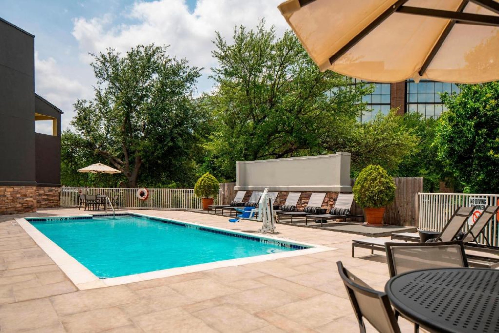Outdoor swimming pool area with lounge chairs, tables, and umbrellas, surrounded by trees and a fence, on a sunny day.