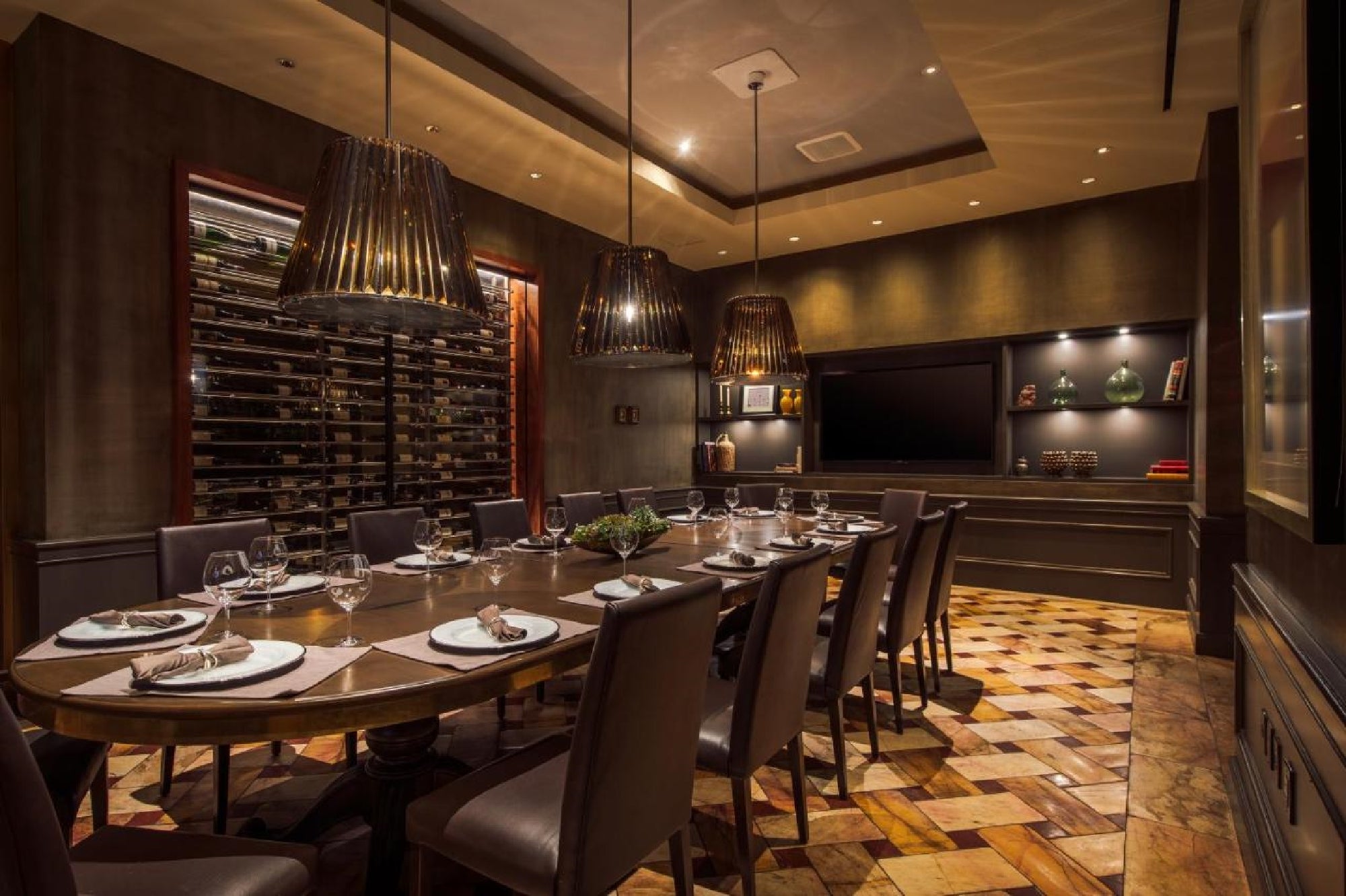 Elegant dining room with a large oval table set with dishes, surrounded by upholstered chairs. Two large pendant lights hang above, and a wine rack is visible on the wall. Wood-patterned floor.