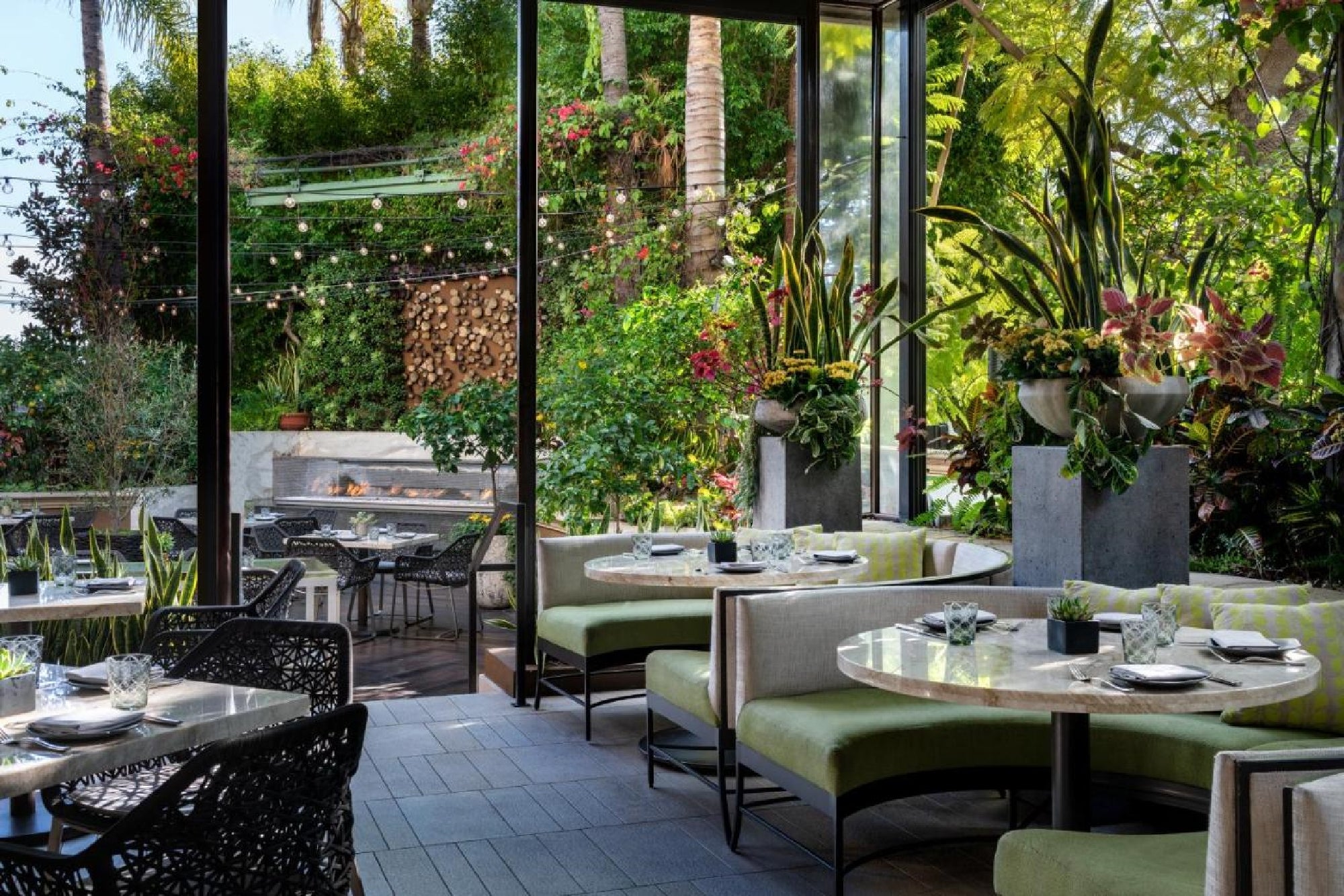 Outdoor restaurant seating with green cushioned booths, round tables set for dining, and lush plants surrounding the area.