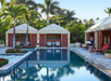 A man walks by a pool holding a towel, with cabanas and palm trees in the background. The setting is sunny and tropical.