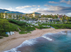 Aerial view of a beachfront resort with palm trees, sandy beach, and ocean waves. Buildings and outdoor amenities are visible in the background.