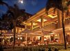 Outdoor restaurant seating area at night with illuminated tables, tropical plants, and large umbrellas. The building features columns and warm lighting under a clear sky with the moon visible.