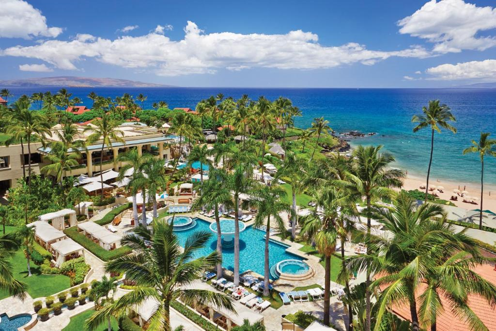 Aerial view of a tropical resort with multiple pools, palm trees, a beach, and ocean in the background under a partly cloudy sky.