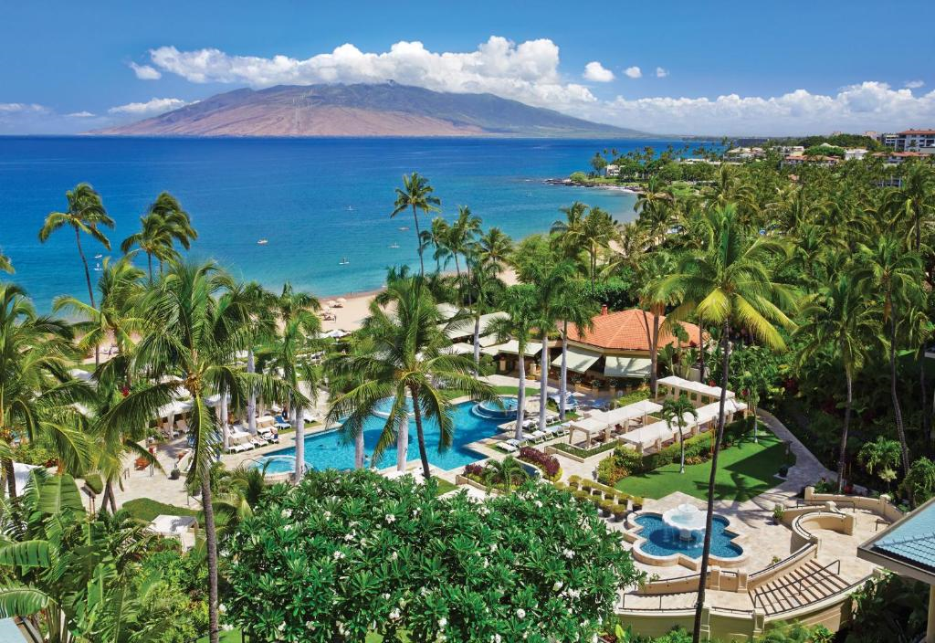 Aerial view of a tropical resort with palm trees, pools, and pathways near a sandy beach. Ocean and mountain range in the background under a blue sky with clouds.
