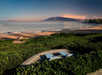 Beachfront view with two empty lounge chairs on grass, overlooking the ocean and distant mountains at sunset.