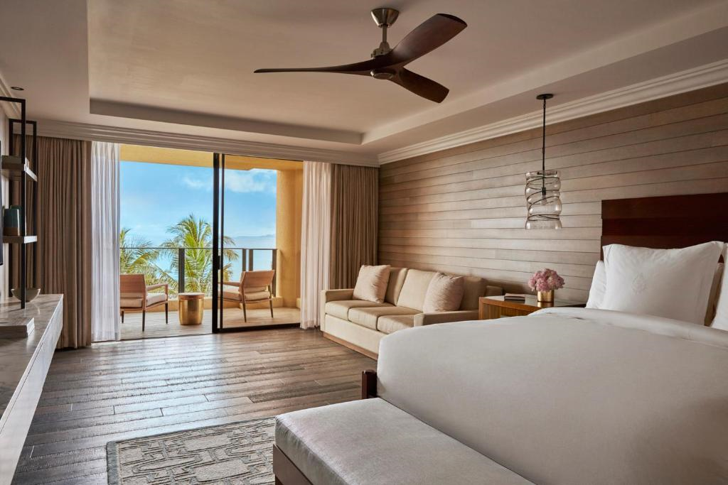 Hotel room with a bed, sofa, and a ceiling fan. Sliding glass doors lead to a balcony with chairs, overlooking a view of the ocean and palm trees. Room features wooden floors and neutral decor.