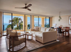 A spacious living room with a beige sectional sofa, two chairs, a rug, and a dining table. Large windows show a view of palm trees and the ocean. A ceiling fan is above.